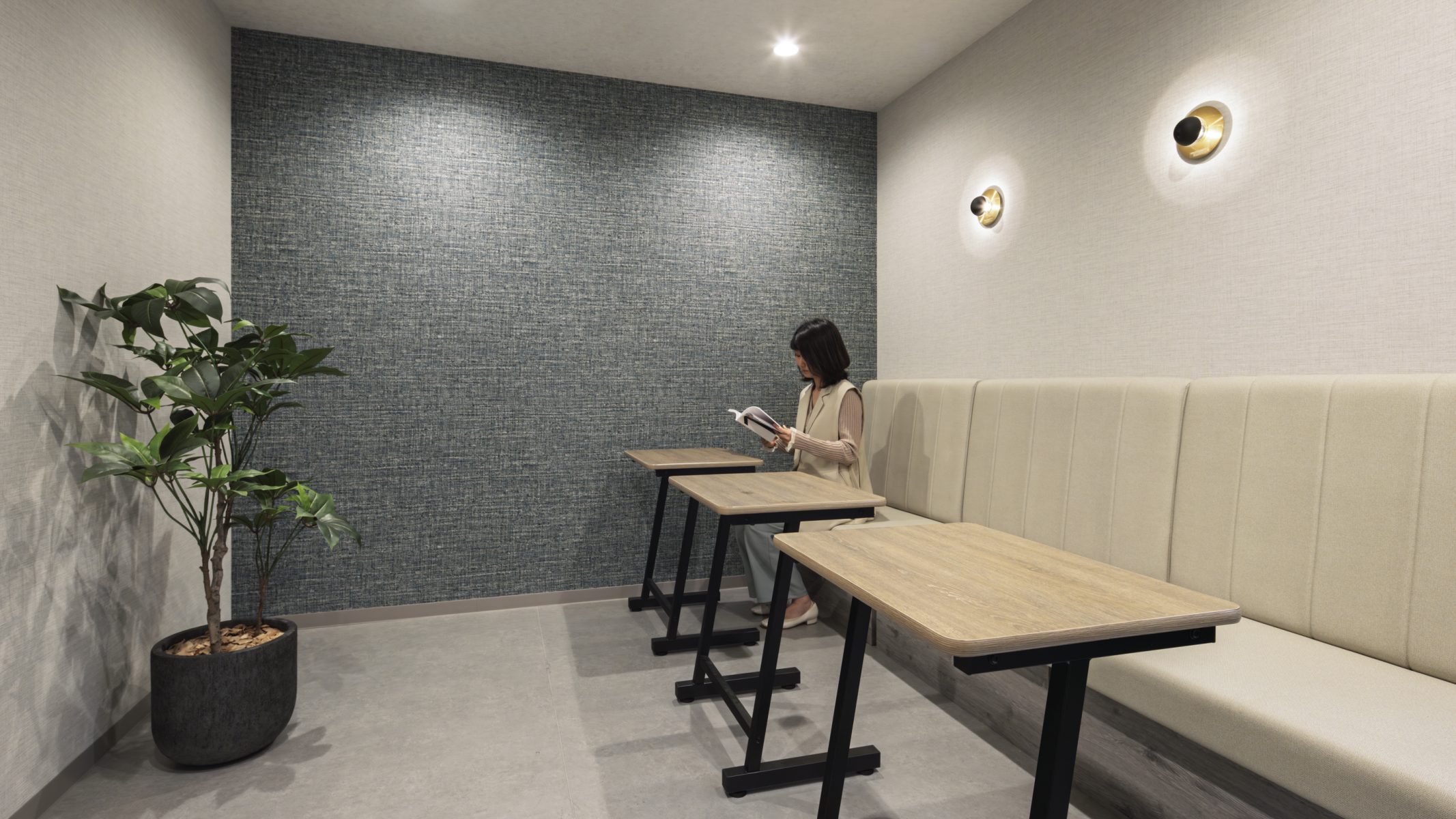 TSネットワーク株式会社 泉大津流通センター ２階休憩室のソファコーナー。グレーのファブリッククロスをアクセントウォールに採用し、ゴールドのブラケット照明とソファ席で一人でくつろげる落ち着いたスペースを設けた。
