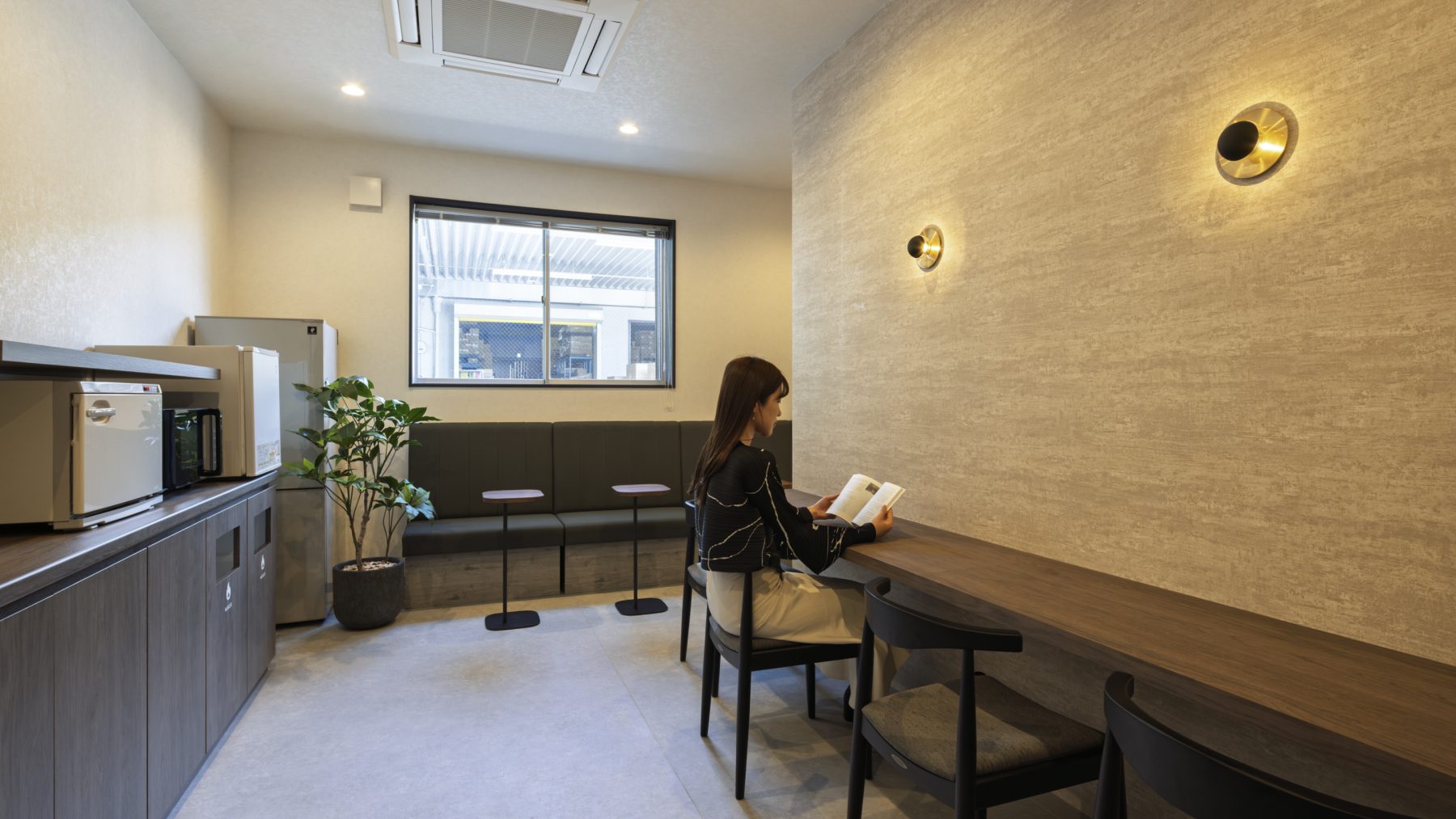 TSネットワーク株式会社 泉大津流通センター １階休憩室。深みのある木目カウンターと石目調クロスのアクセントウォール、ブラケット照明を組み合わせ、１階全体のシックなデザインに調和した落ち着きのある休憩空間。