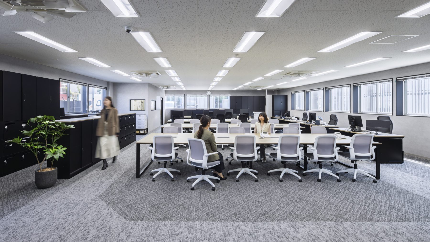 TSネットワーク株式会社 泉大津流通センター １階執務室。グレー系のカーペットとブラックの什器でまとめたシックな執務空間。広い窓から自然光が差し込み、集中しやすい落ち着いた環境を実現している。